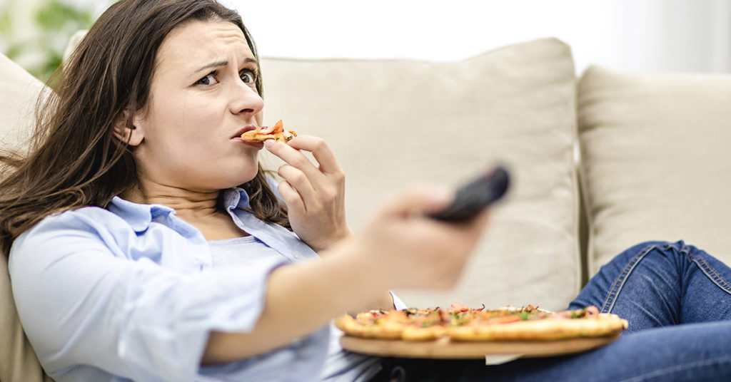 woman eating pizza while on couch using TV remote 