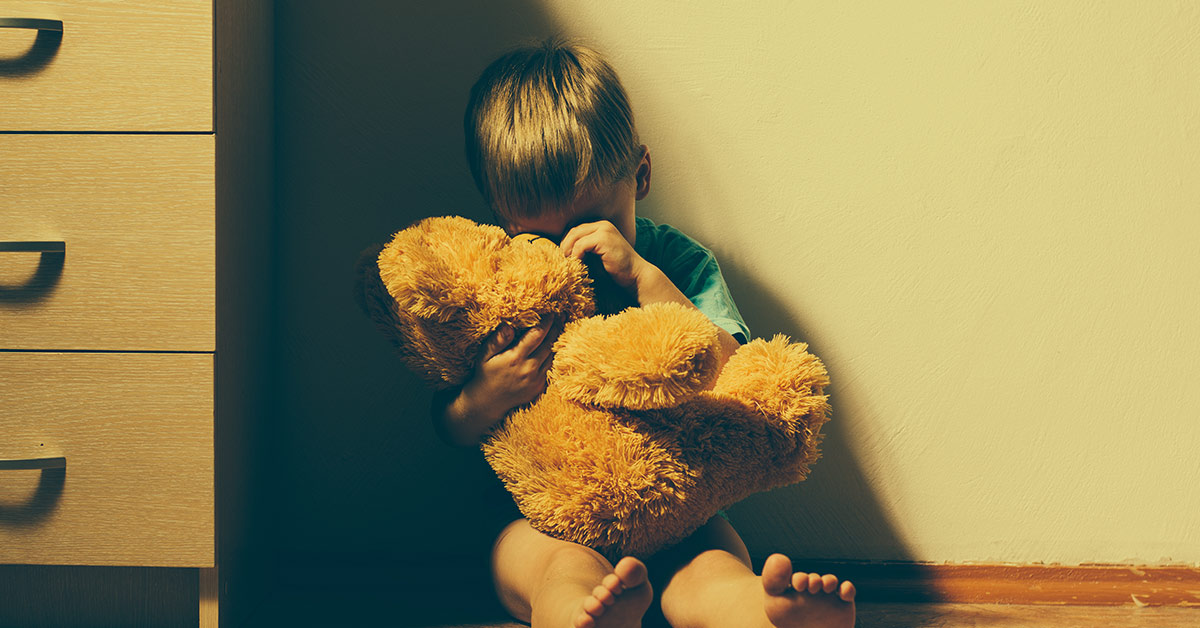sad child sitting against a wall holding a stuffed bear