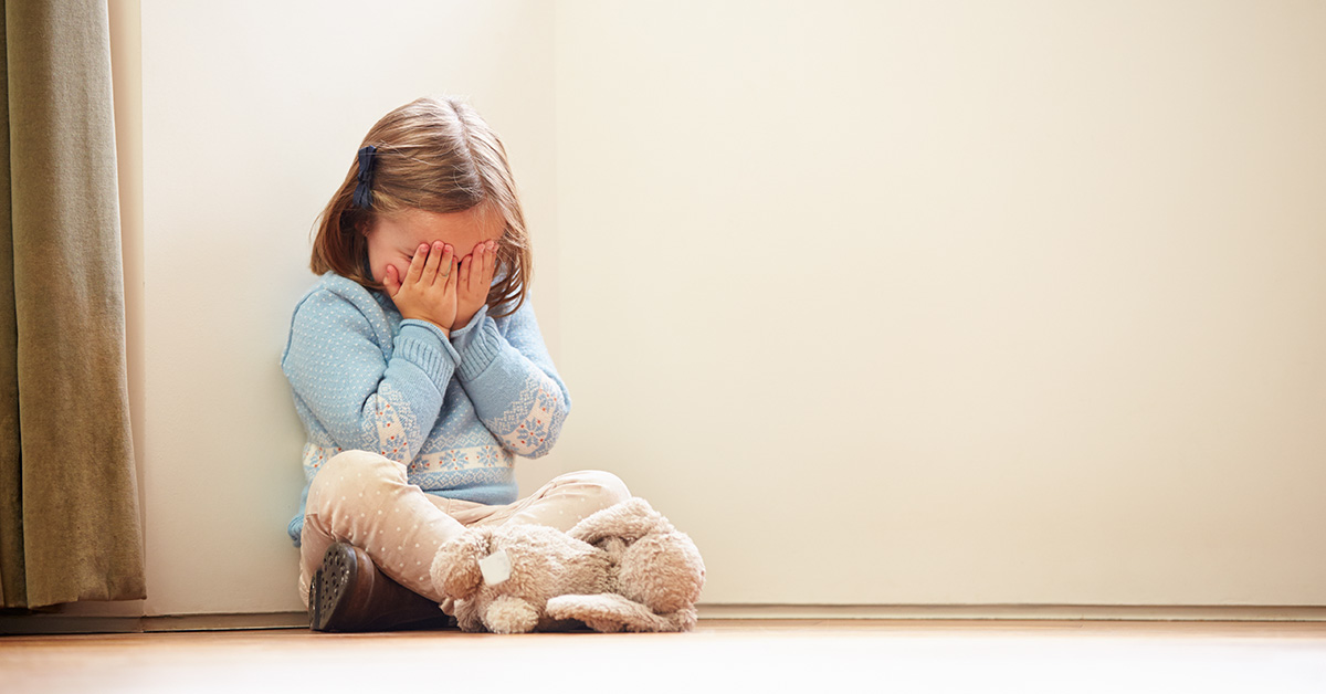toddler girl sitting against a wall crying with hands over eyes. Her stuffed animal lays at her feet