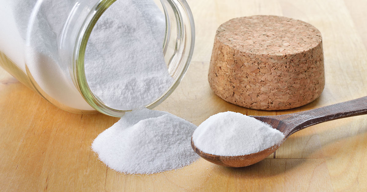 jar of baking soda on its side with some pouring out onto a counter, a cork lid to the right and a spoon full of baking soda