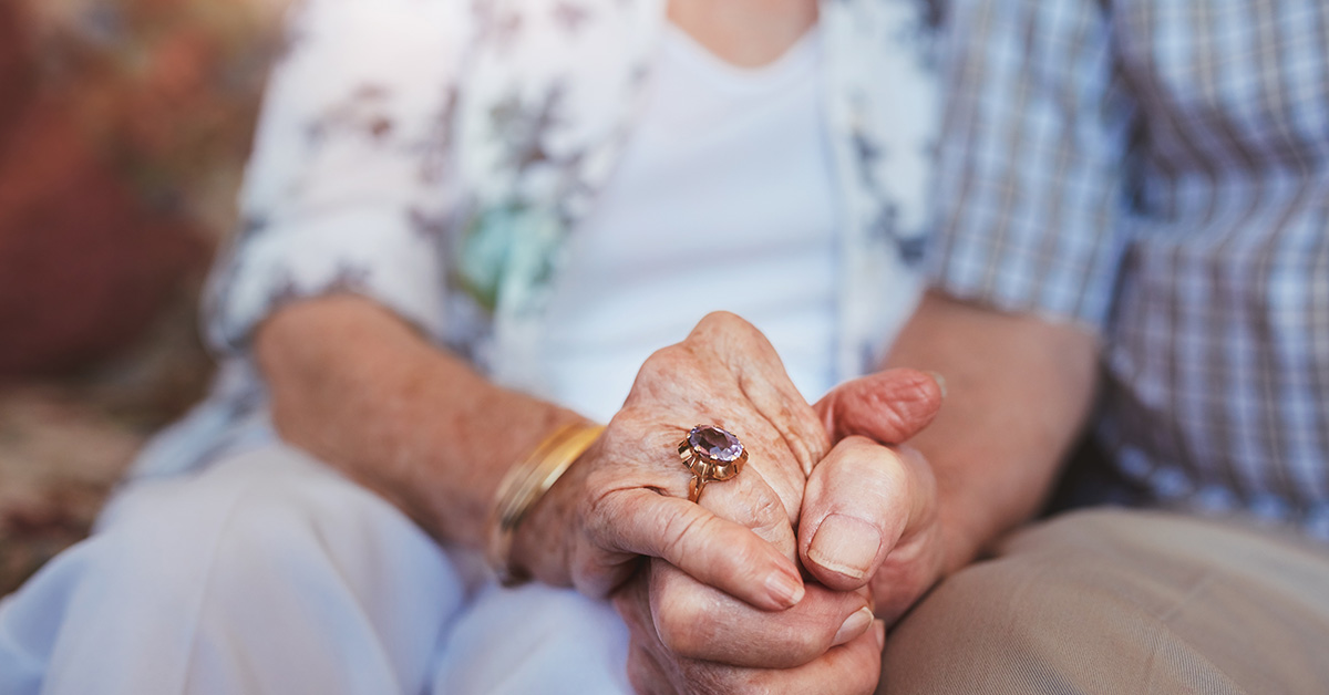 elderly couple holding hands. One hand has a ring with a large purple jewel