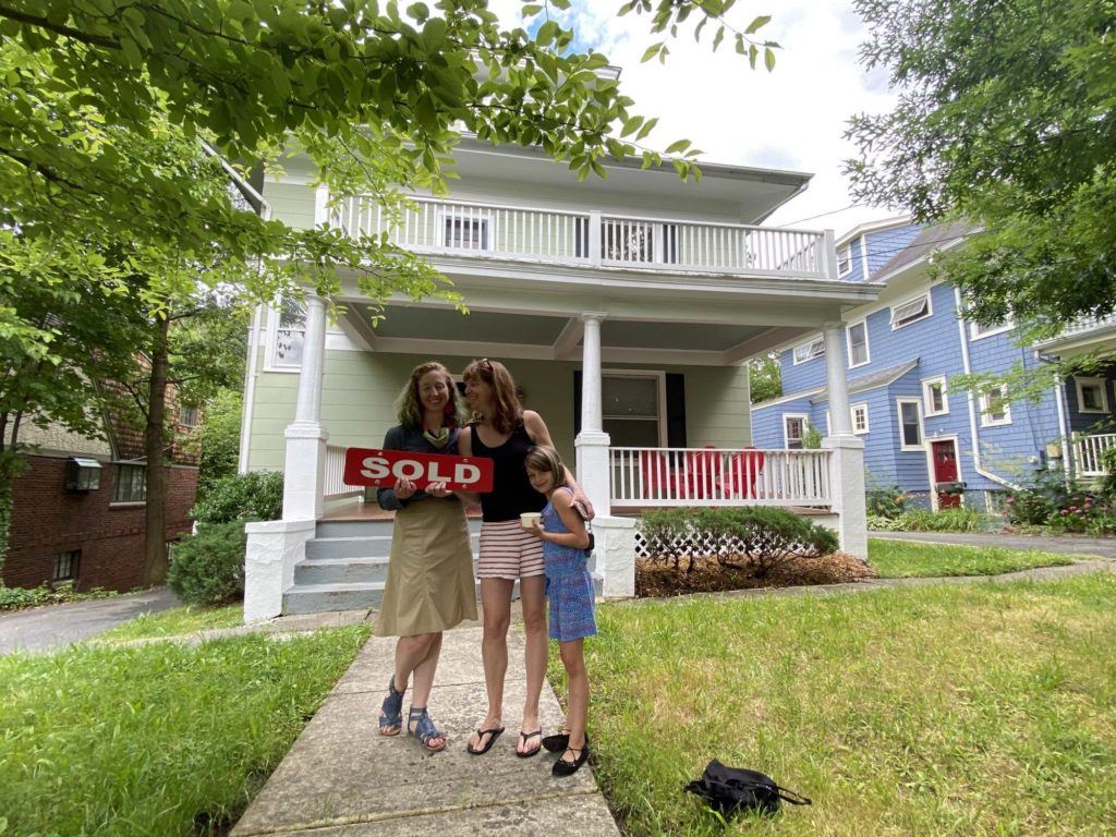 two single mothers and one of their daughters standing outside their newly purchased home while holding a "sold" sign