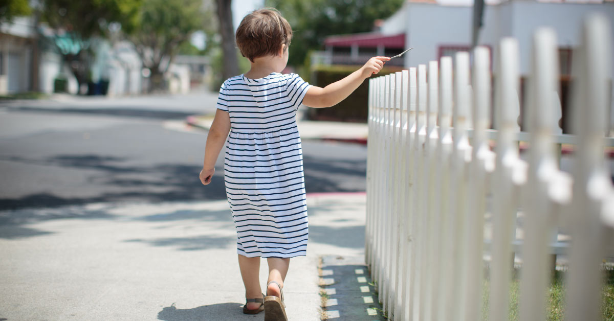 little girl seen from behind walking down a sidewalk running a stick along a white picket fence