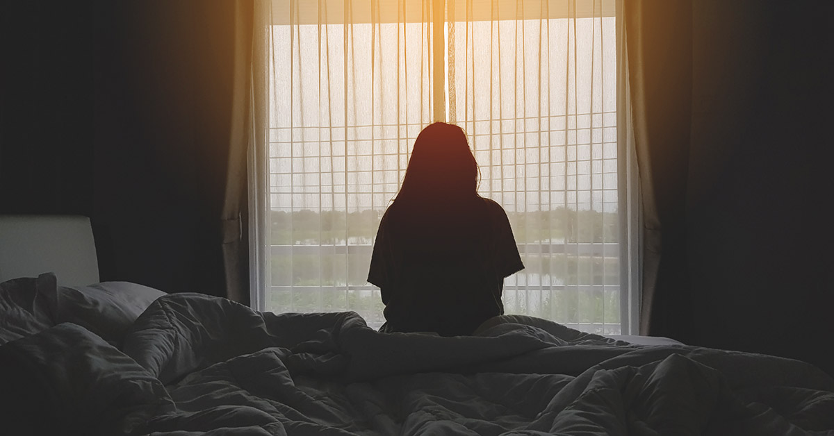 woman sitting on side of bed looking out a window at sunset