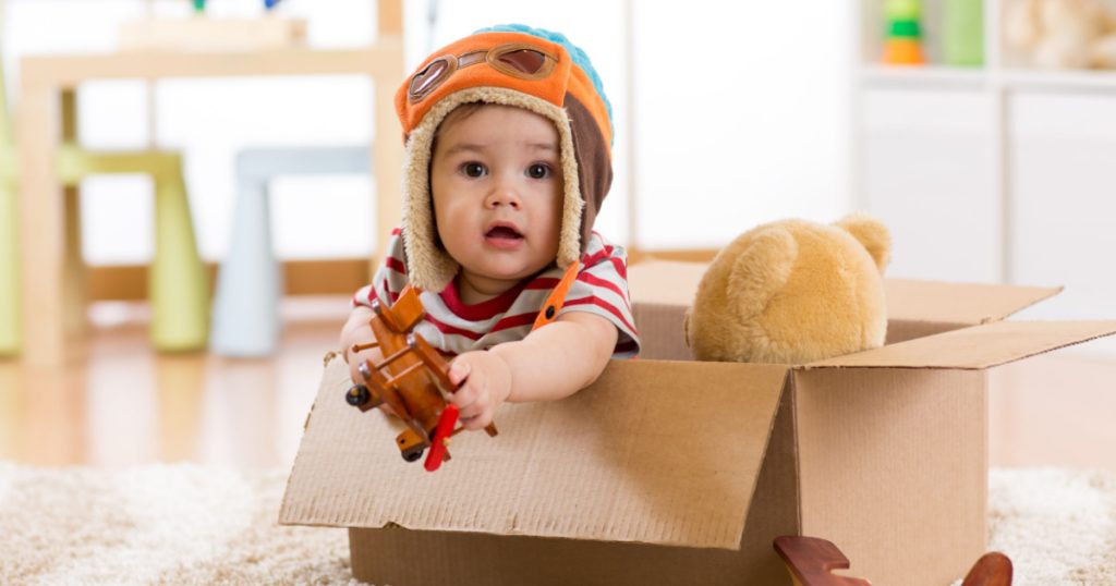 baby boy with teddy bear toy and planes plays in cardboard box

