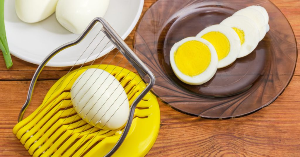 Egg slicer with egg in it closeup, boiled whole peeled and partly sliced eggs in different sausers on an old rustic table at selective focus
