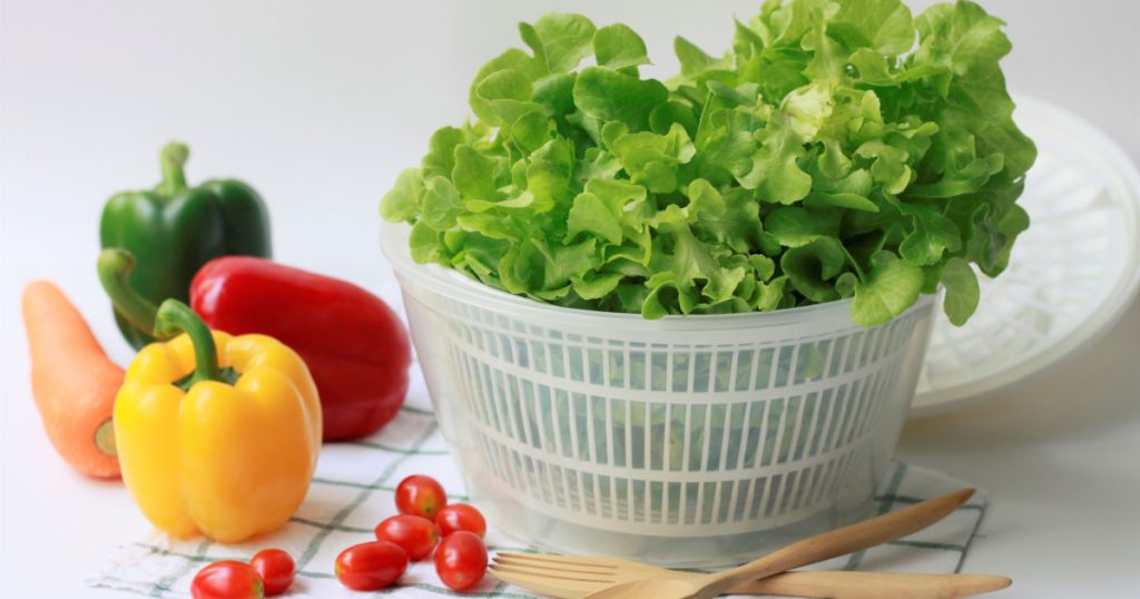 Fresh green oak vegetables in white plastic salad spinner or bowl preparing for mix healthy salad with tomatoes, bell peppers and carrot with fork and spoon on white background