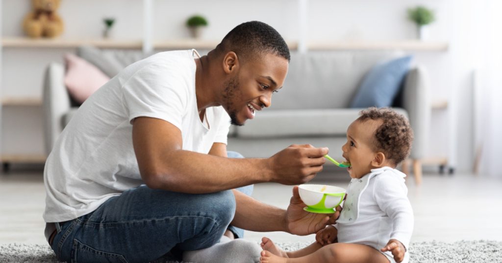 Father's Care. Loving Black Dad Feeding His Cute Baby Son From Spoon At Home, Young African American Daddy Giving Healthy Food To His Little Toddler Child While They Relaxing Together In Living Room