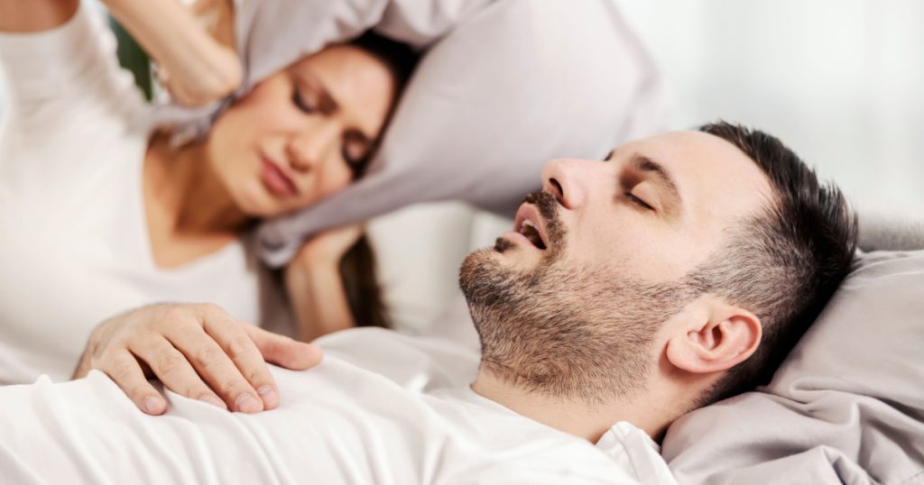 A man snoring and sleeping in the bed while woman is annoyed
