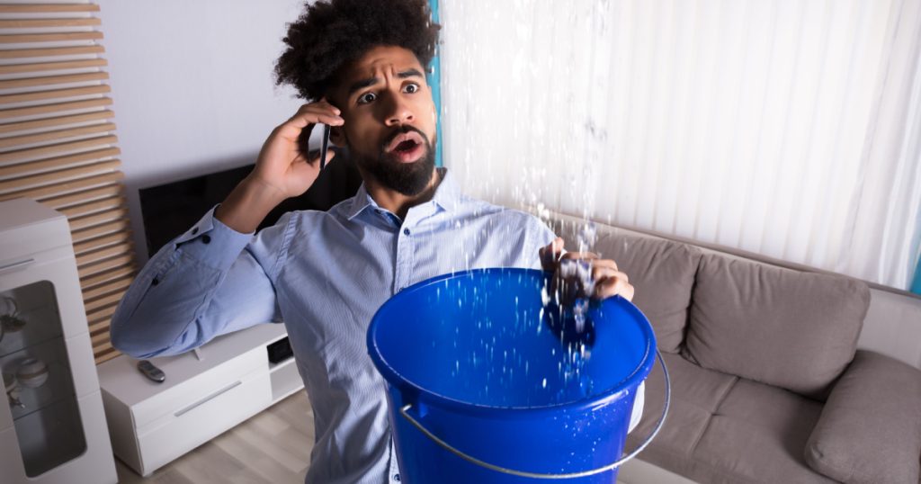 Worried Young Man Calling Plumber While Leakage Water Falling Into Bucket At Home
