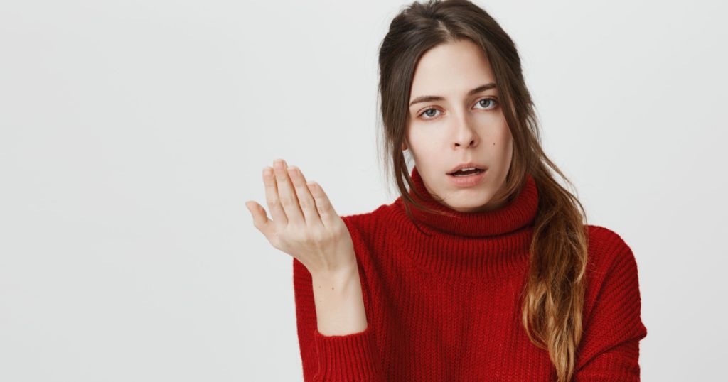 Irritated young female with dark hair in ponytail has displeased face expression, gestures actively, expresses negativeness, isolated against gray background. Girl asking so what, who cares