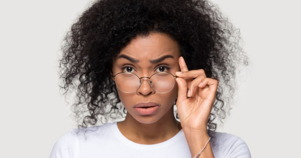 Studio portrait of shocked african young woman teacher wearing white t-shirt lowering glasses looking at camera feels confused surprised received amazing stunned news posing isolate on grey background
