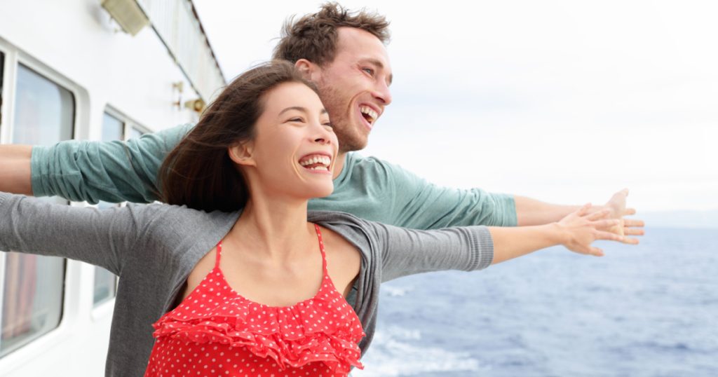 Romantic couple having fun laughing in funny pose on cruise ship boat. Smiling happy man and woman on travel vacation holidays on open ocean sea.
