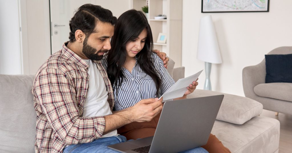 Young indian couple reading paper bills paying loan bank debt online together on computer, calculating taxes, income, making payments, planning family budget money finances using laptop at home.
