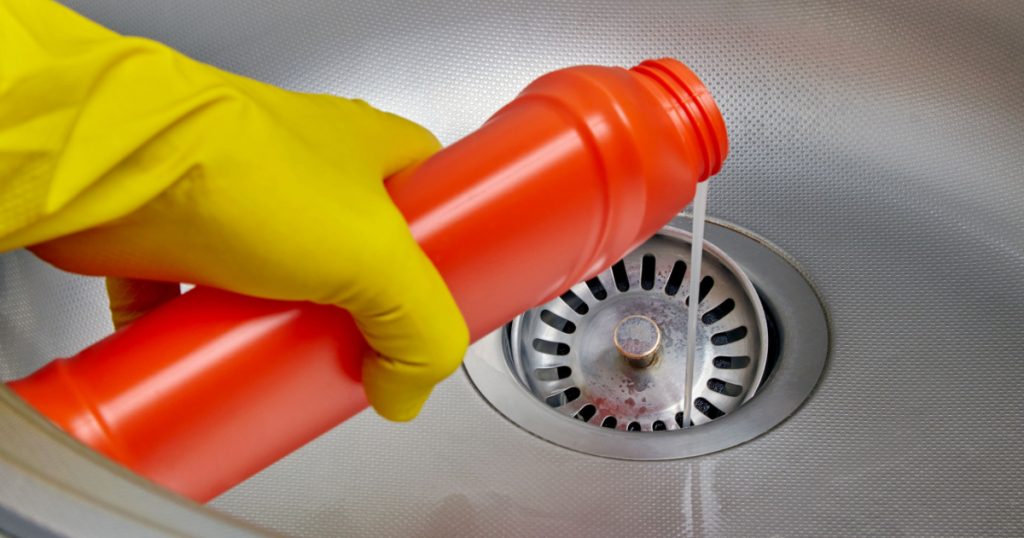 Person's hand in a yellow rubber glove pours pipe cleaner down the drain of a metal kitchen sink