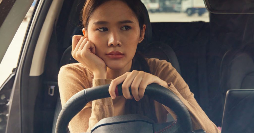 Woman driving alone negative emotions : Asian woman with her hands on her chin feeling overwhelmed tired stressed out at work overthinking and absent minded in the car while driving distracted driving