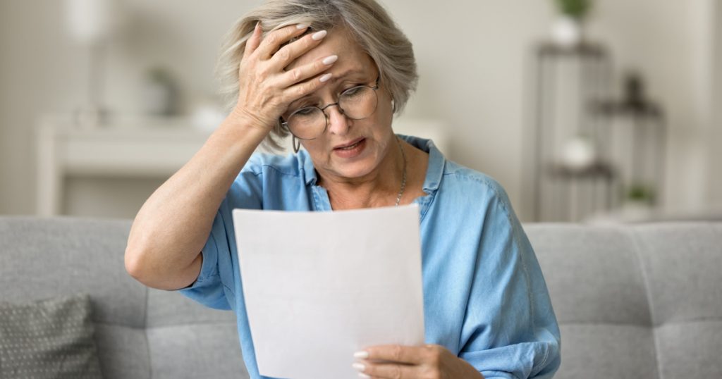 Stressed frustrated older woman getting bad news from paper letter, reading document at home, touching head in despair, panic attack, sitting on couch, receiving concerning medical result

