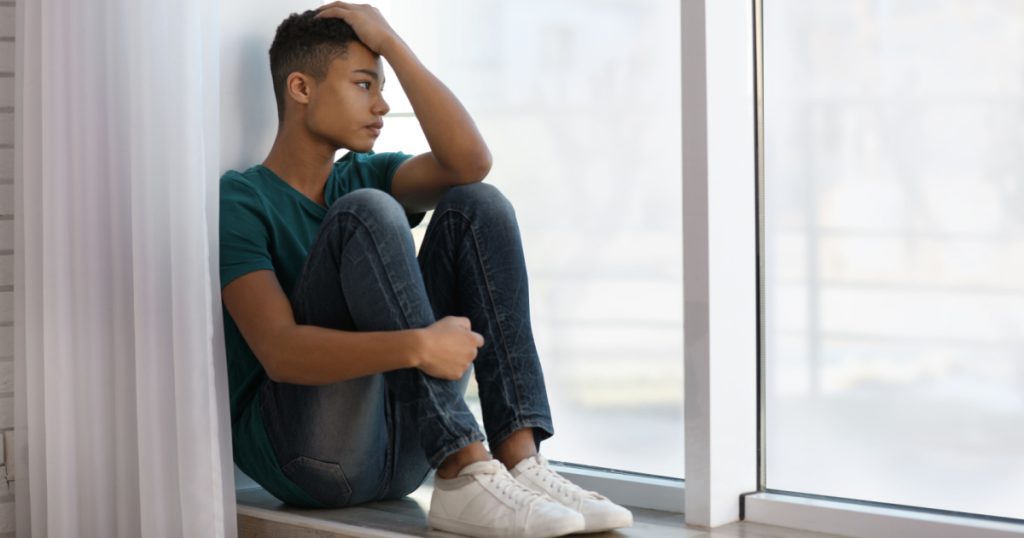 Upset African-American teenage boy sitting alone near window
