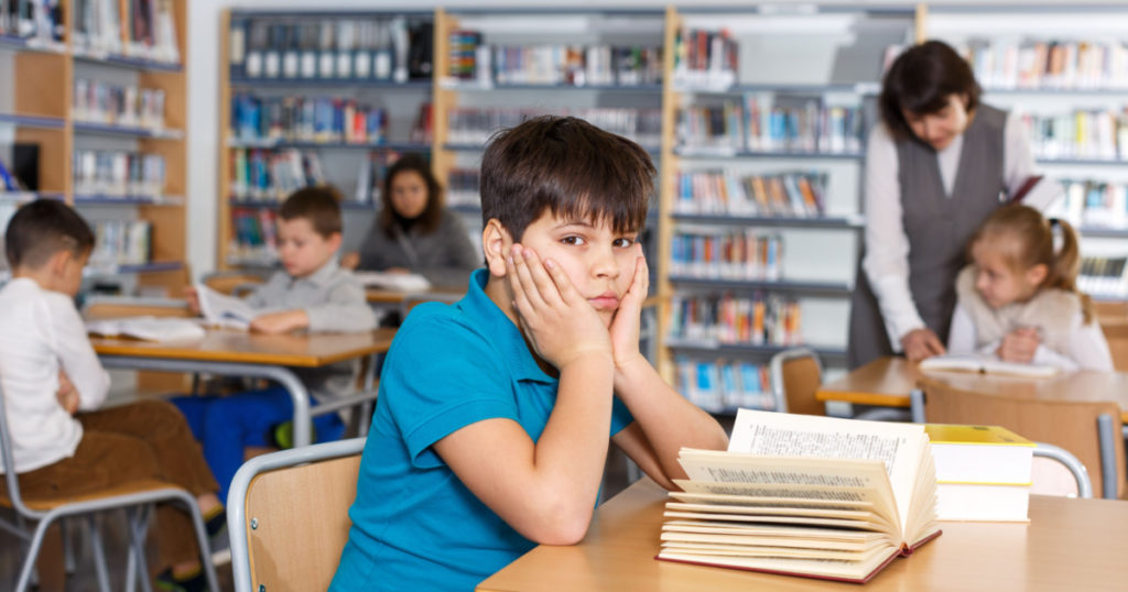 Portrait of upset tween boy reading in school library on background with other students and teacher
