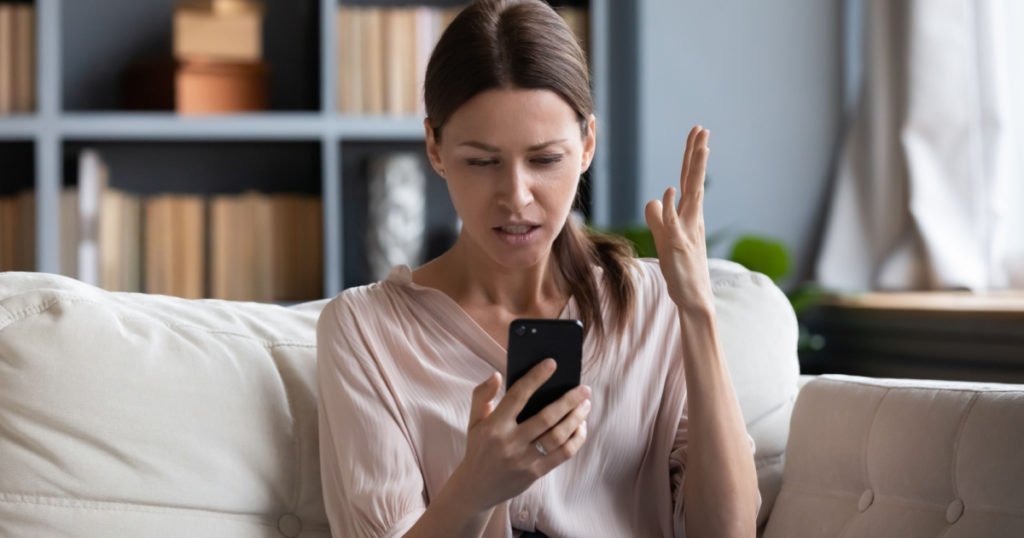 Confused angry woman having problem with phone, sitting on couch at home, unhappy young female looking at screen, dissatisfied by discharged or broken smartphone, reading bad news in message
