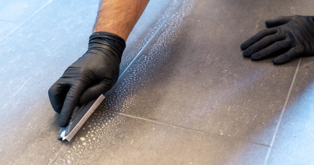A close up of a professional cleaner cleaning grout with a brush blade and foamy soap on a gray tiled bathroom floor
