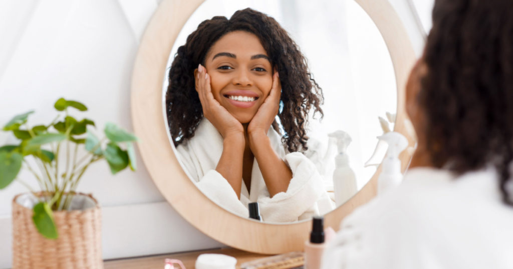 Self-Care. Beautiful African American Woman In Bathrobe Ready To Apply Makeup, Reflecting In Mirror Of Toilette Dresser Table, Looking At Camera
