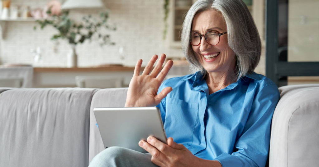 Happy 60s older mature middle aged adult woman waving hand holding digital tablet computer video conference calling by social distance virtual family online chat meeting sitting on couch at home.
