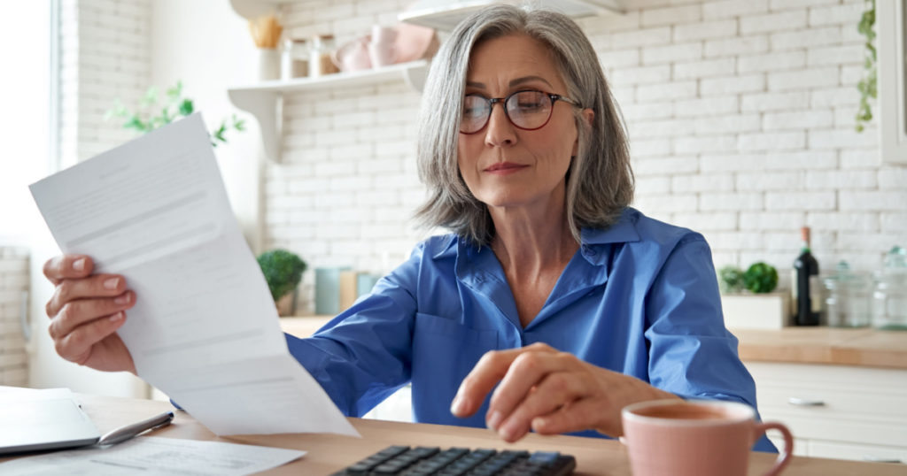 Senior mature business woman holding paper bill using calculator, old lady managing account finance, calculating money budget tax, planning banking loan debt pension payment sit at home kitchen table.
