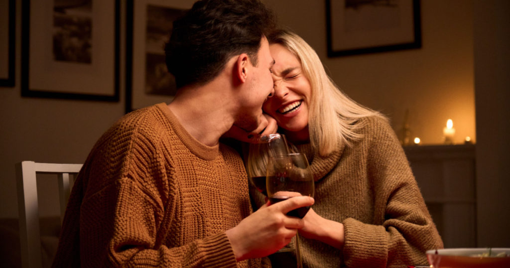 Happy young couple in love hugging, laughing, drinking wine, enjoying talking, having fun together celebrating Valentines day dining at home, having romantic dinner date with candles sitting at table.
