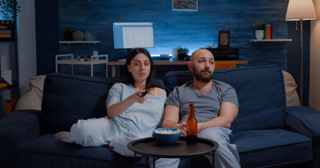 Young couple spending evening together dressed in pajamas arguing for remote control, wife switching channels changing tv programs trying to relax on sofa together eating popcorn, drinking beer
