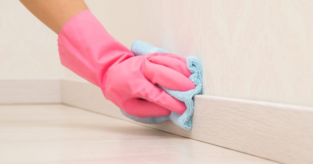Young adult woman hand in pink rubber protective glove using blue dry rag and wiping light wooden baseboard surface in room at home. Closeup.
