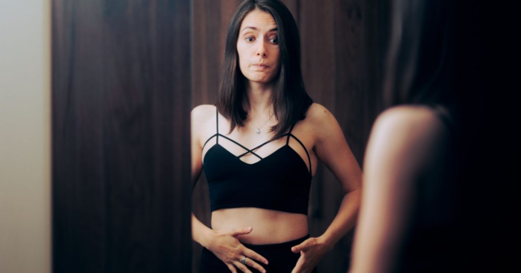 Insecure Self-conscious Woman Checking her Body in the Mirror. Girl feeling bloated looking at her stomach in mirror reflection
