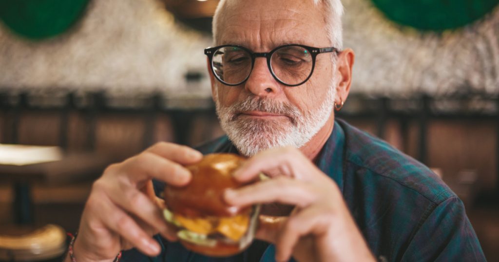 Senior gray-haired man in glasses with a beard is eating an appetizing burger. Lunch break, rest in the pub.
