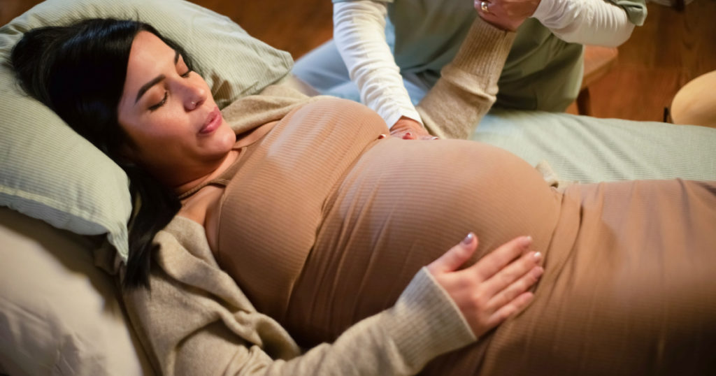Cropped image of pregnant woman and helpful midwife at home. Woman in casual clothes lying on bed, midwife holding hand. Pregnancy, medicine, home birth concept
