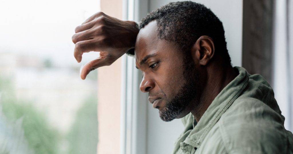 Unhappy frustrated adult african american guy in casual suffering from depression and bad news near window in home interior. Health problems, stress from self-isolation during covid-19 quarantine
