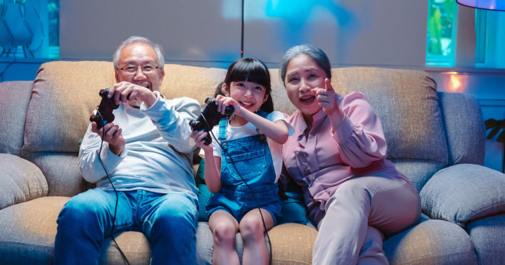 Asian happy family play time. little girl with grandparents together playing video games use joystick control enjoy laughing fun in living room home at night. Happy child happiness lifestyle concept.
