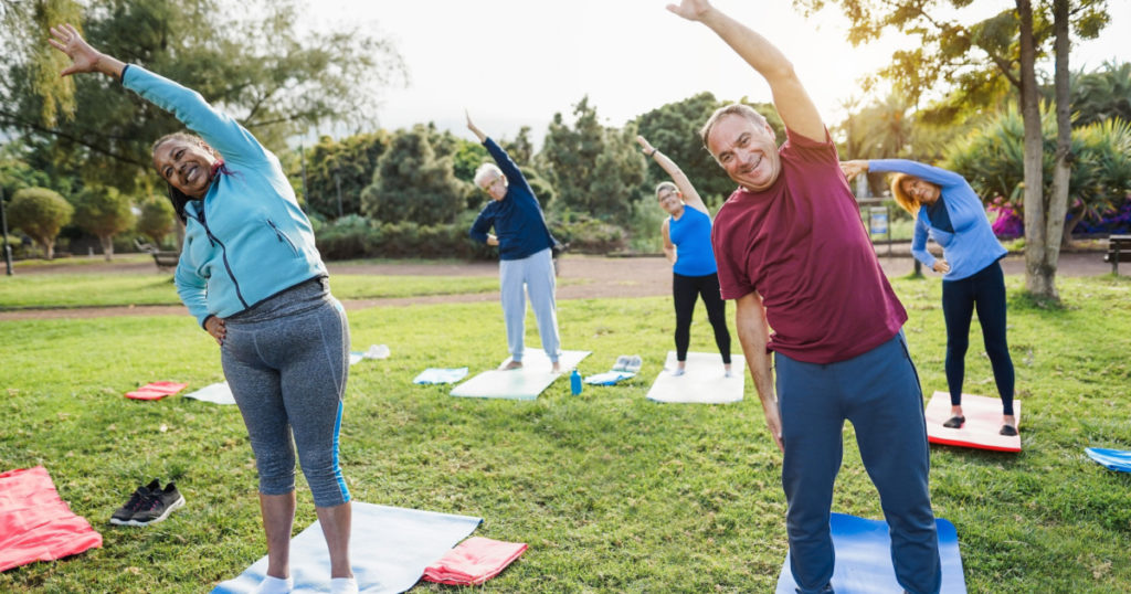 Multiracial senior people doing stretching workout exercises outdoor with city park in background - Healthy lifestyle and joyful elderly lifestyle concept - Focus on right man face
