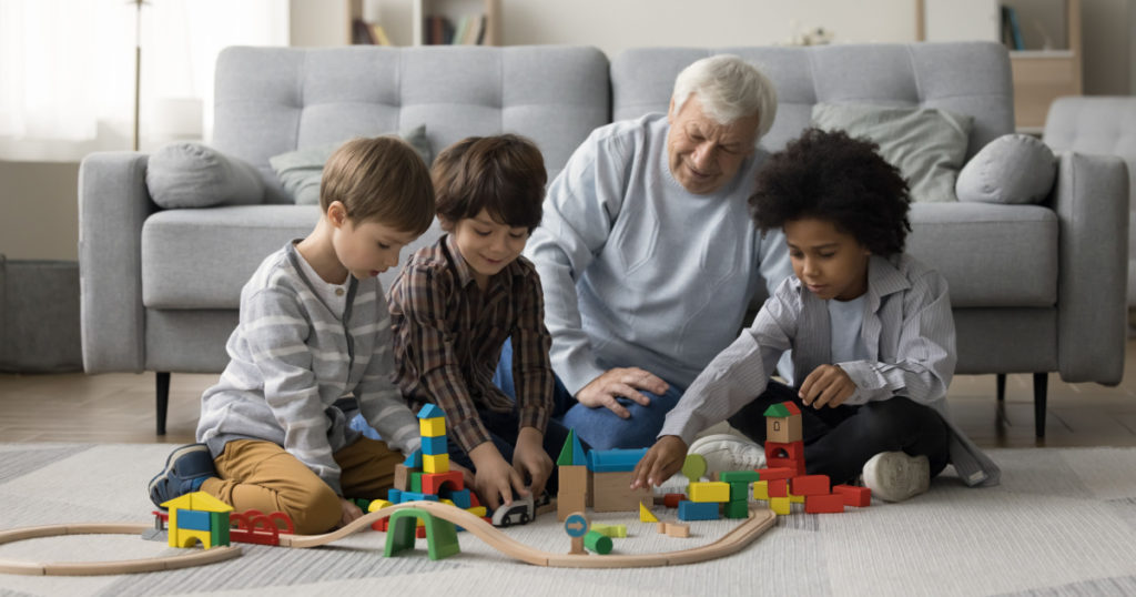 Elderly grandpa watching three multiethnic little grandsons playing game with toy construction blocks on warm floor, building city towers model together, enjoying family playtime, teamwork
