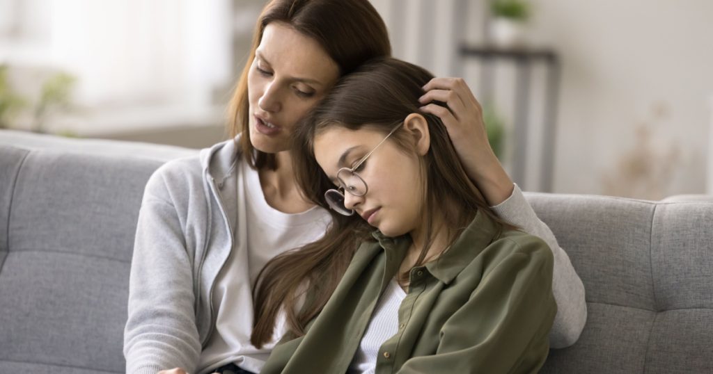 Compassionate young mother comforting depressed sad teen girl, hugging daughter with love, care, speaking, giving sympathy, advice, support, holding kids hand, sitting on home couch

