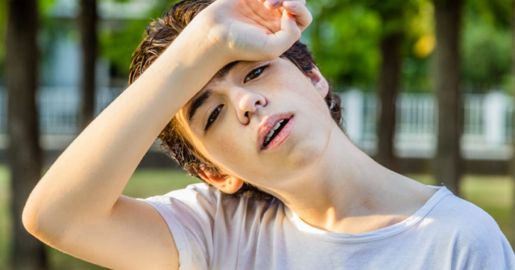 Tired teenager with acne skin holds his head protecting eyes from sunlight with arm on front while showing braces with trees in the background
