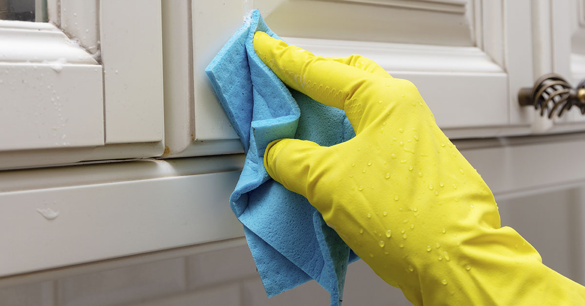 hand with yellow cleaning gloves scrubbing a kitchen cabinet with a blue cloth