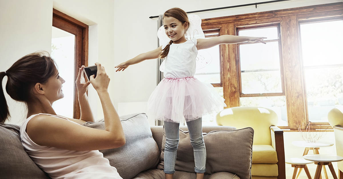 mother taking picture of daughter in a tutu standing on couch