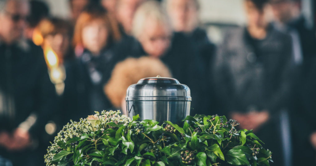 A metal urn with ashes of a dead person on a funeral, with people mourning in the background on a memorial service. Sad grieving moment at the end of a life. Last farewell to a person in an urn.
