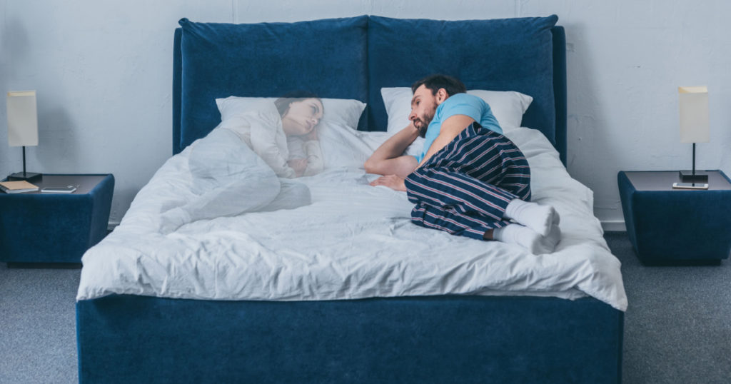 depressed man lying in bed and looking at wife ghost
