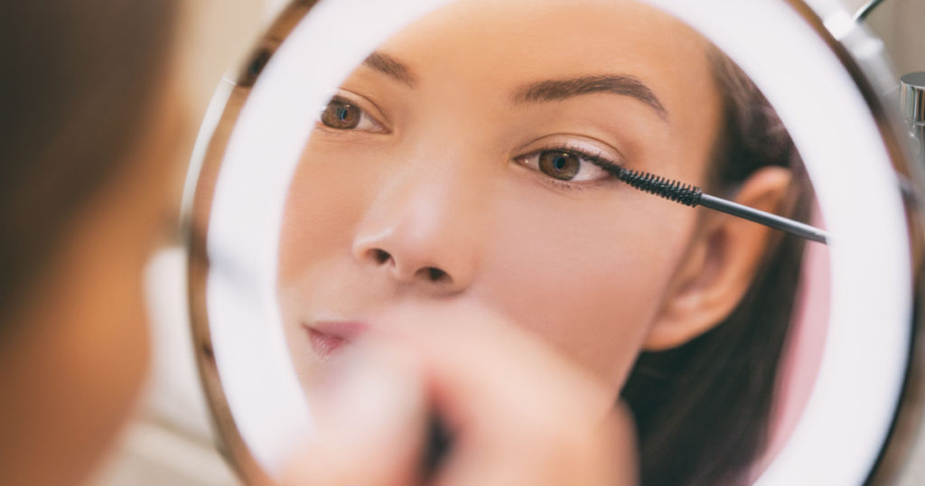 Woman applying make up beauty product putting mascara in ring lighted round makeup mirror at home bathroom morning routine. Beautiful Asian lady getting ready applying eye make-up with brush.
