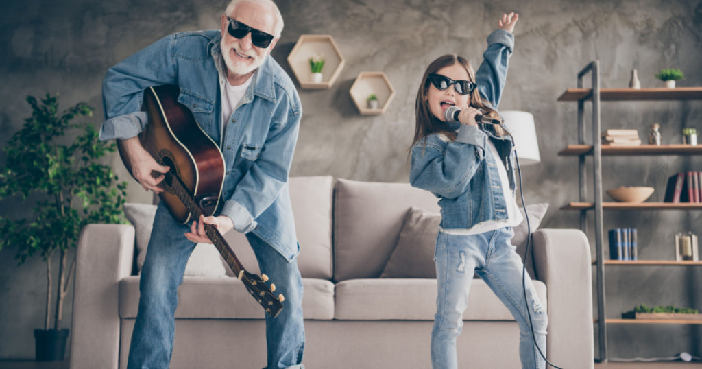 Photo of two people funky grandpa play guitar small nice granddaughter mic singing cool style sun specs denim clothes repetition school concert stay home quarantine living room indoors
