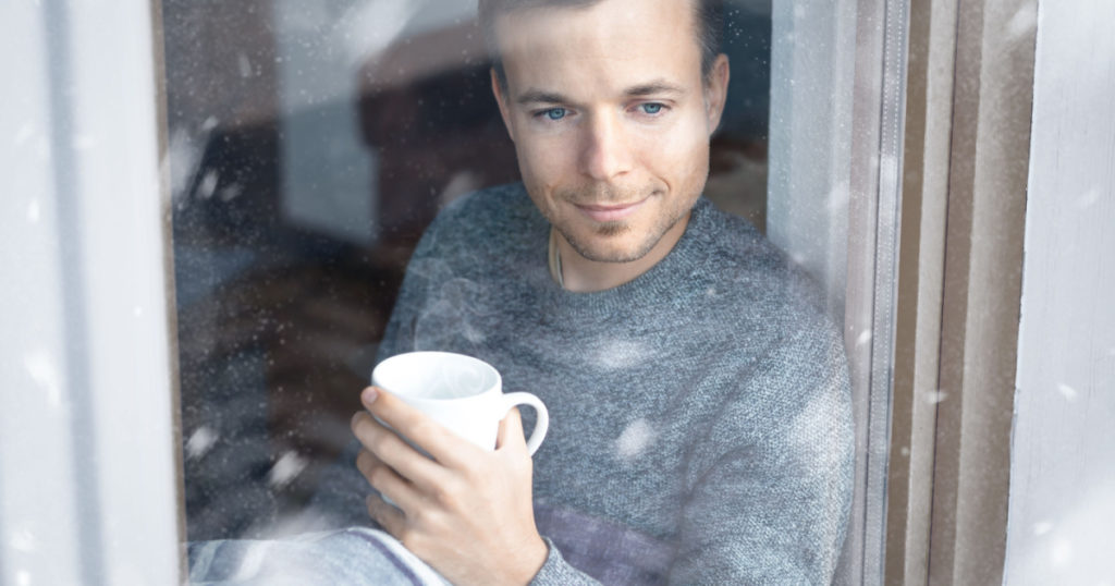 Man with a cup in his hand is sitting at a window during winter
