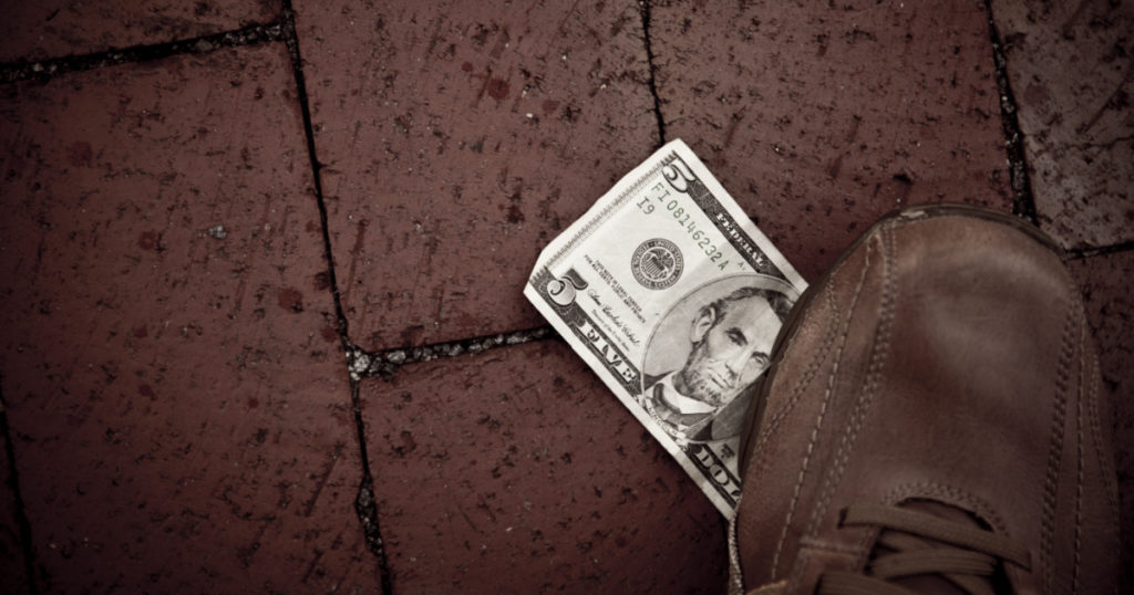 A person finds a US five dollar bill at the street and he uses his foot to step on it.
