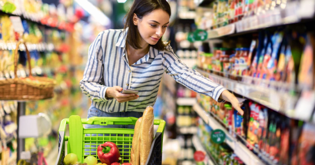 Portrait Of Millennial Lady Holding And Using Smartphone Buying Food Groceries Walking In Supermarket With Trolley Cart. Female Customer Shopping With Checklist, Taking Products From Shelf At The Shop
