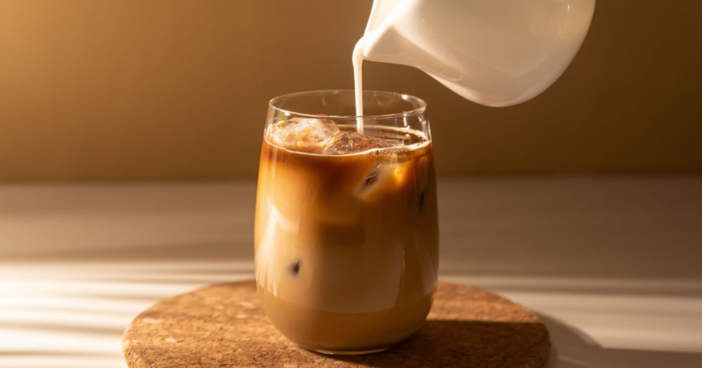 Milk cream is poured into a iced cold brew coffee. Coffee cold cocktail drink with ice and milk in morning sun light.
